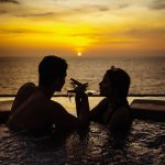A couple relaxes in a pool, sipping wine while watching a beautiful sunset over the ocean.
