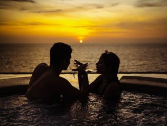 A couple relaxes in a pool, sipping wine while watching a beautiful sunset over the ocean.