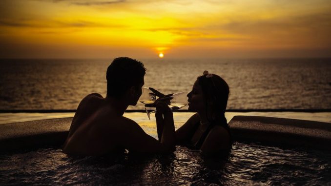 A couple relaxes in a pool, sipping wine while watching a beautiful sunset over the ocean.