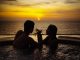 A couple relaxes in a pool, sipping wine while watching a beautiful sunset over the ocean.