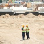 Two workers discussing plans on a sandy construction site, wearing safety gear.