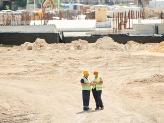 Two workers discussing plans on a sandy construction site, wearing safety gear.