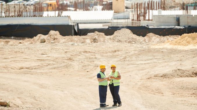Two workers discussing plans on a sandy construction site, wearing safety gear.