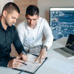 Two businessmen analyzing financial charts on digital devices in a modern office setting.