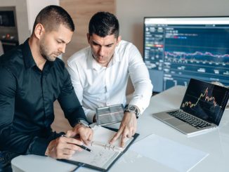 Two businessmen analyzing financial charts on digital devices in a modern office setting.