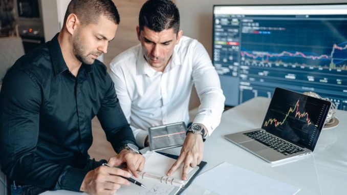 Two businessmen analyzing financial charts on digital devices in a modern office setting.